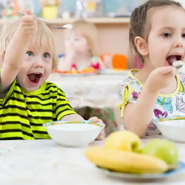 Two children in a childcare centre sitting at a table eating a meal with bowls and fruits.