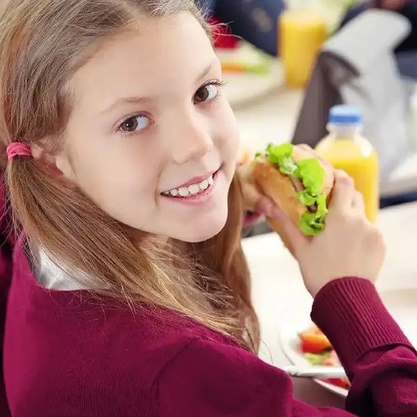 Young girl eating school lunch. She is holding a sandwich with a smile, sitting at a table with a bottle of juice in the background.