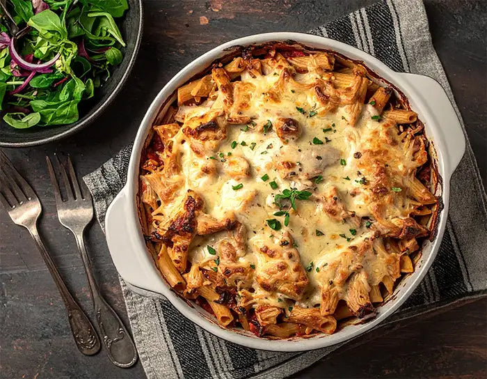 Baked chicken pasta dish with cheese in a white dish on a wooden table with salad and utensils.