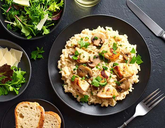 Risotto with mushrooms on a black plate, surrounded by salad, bread, and a glass of white wine.