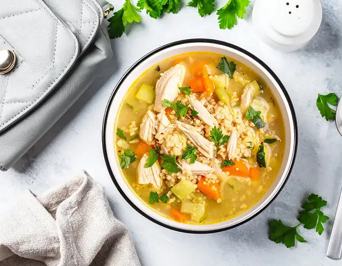 Bowl of chicken soup with vegetables on a light surface with a handbag.