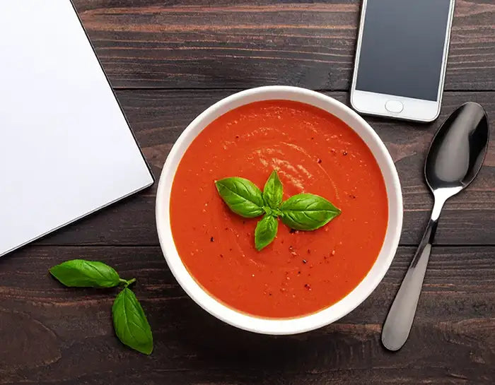 Bowl of tomato soup with basil leaves on a wooden table with a phone and notebook.