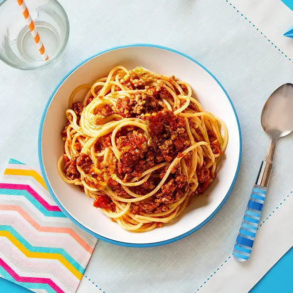 Plate of spaghetti bolognese on a table with a glass of water and colorful napkin.