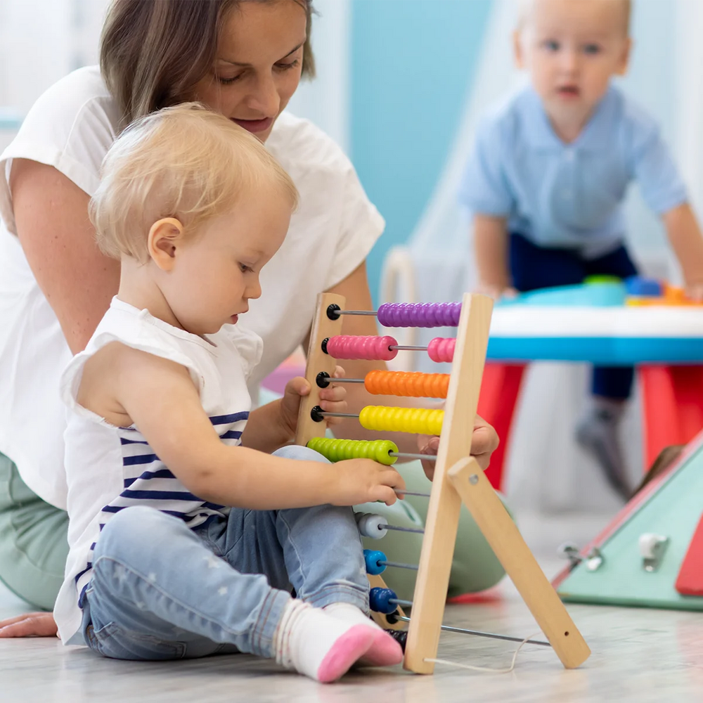 A scene pictured in a childcare centre of a The Little Gourmet customer. A young women childcare educator playing with an abacus alongside a cute toddler. Another toddler is in the background playing with a toy.