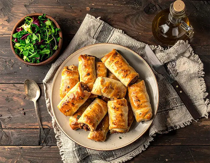 Platter of vegetable sausage rolls on a wooden table with salad and oil bottle.