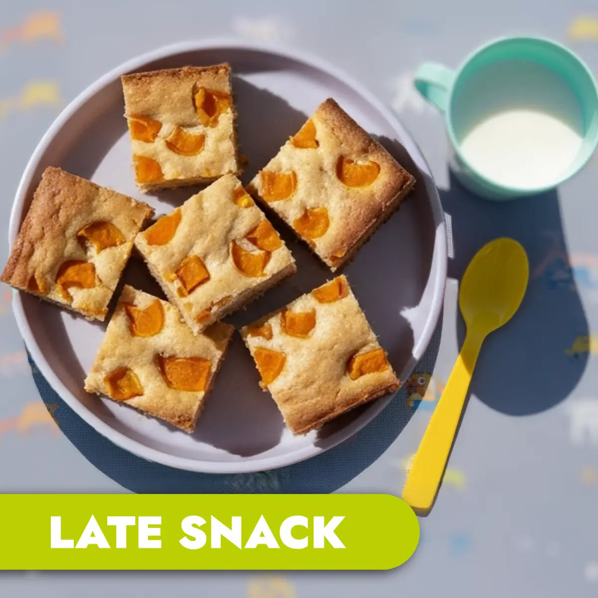 Plate of square cake bars with apricot pieces, a green cup of milk, and yellow spoon on a light blue background.